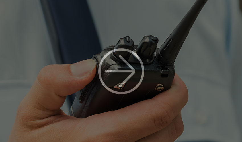 Person hält ein Walkie-Talkie und konzentriert sich auf die Bedienelemente. Der unscharfe Hintergrund betont die Kommunikation in einem professionellen Umfeld.