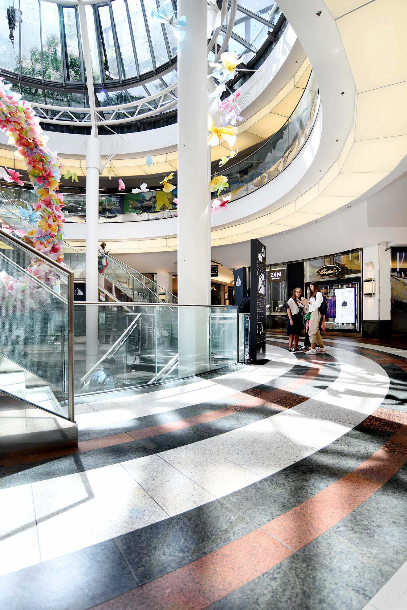 The interior of a vibrant shopping center with colorful paper decorations and sunlight creating patterns on the polished floor and glass railing.