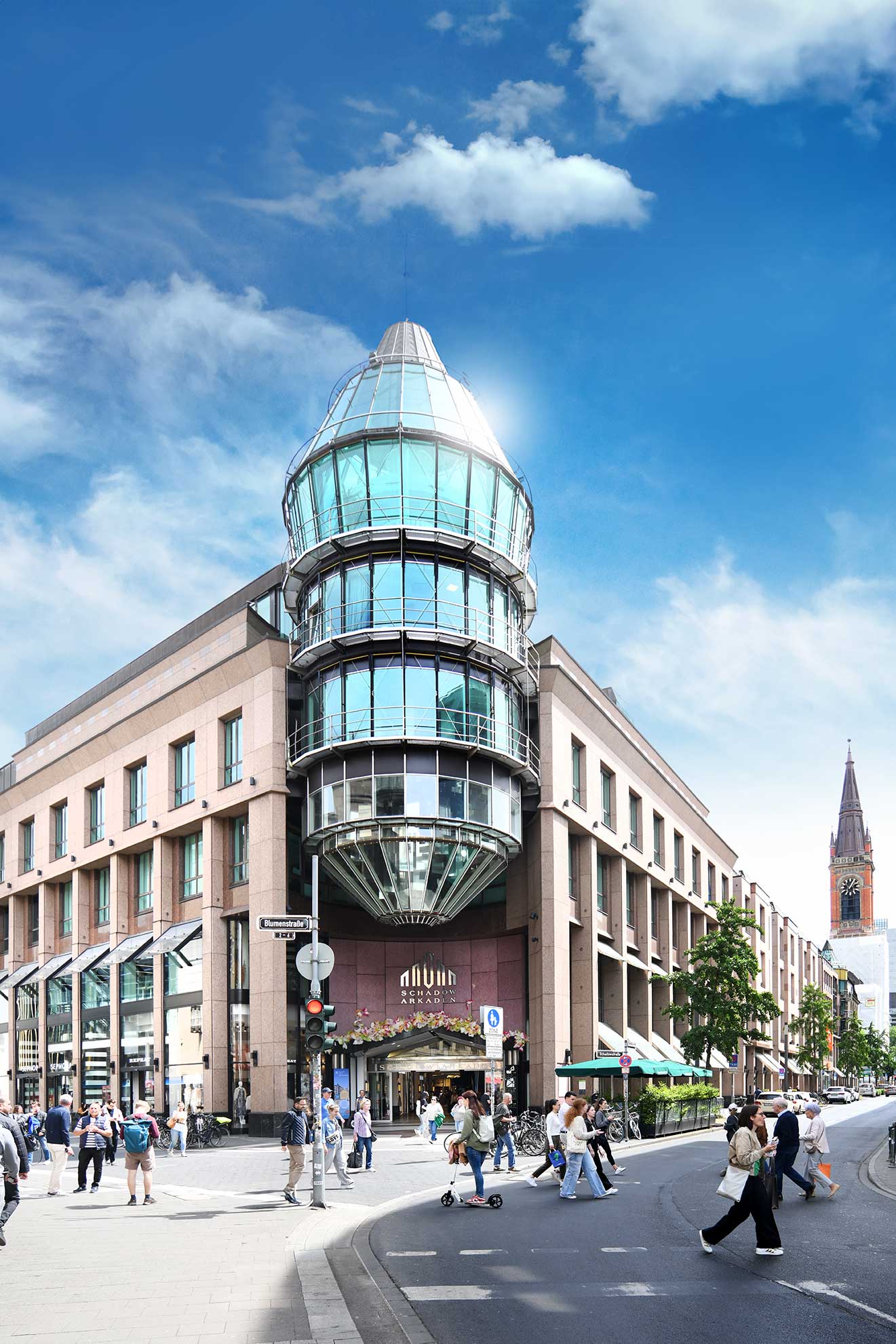 The Schadow Arkaden from the outside, a modern shopping center made of glass and brick with a distinctive tower, bustling with many pedestrians on a sunny day in an urban setting.