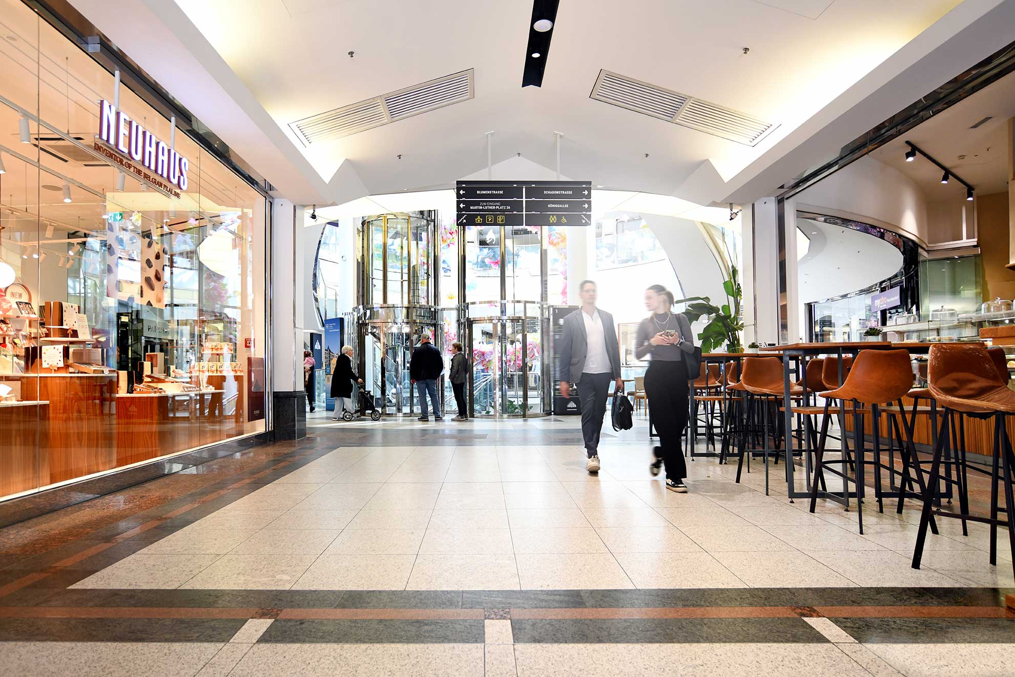 The interior of a modern shopping center with a Neuhaus store, an elevator, and customers walking among stylish decor and seating areas.