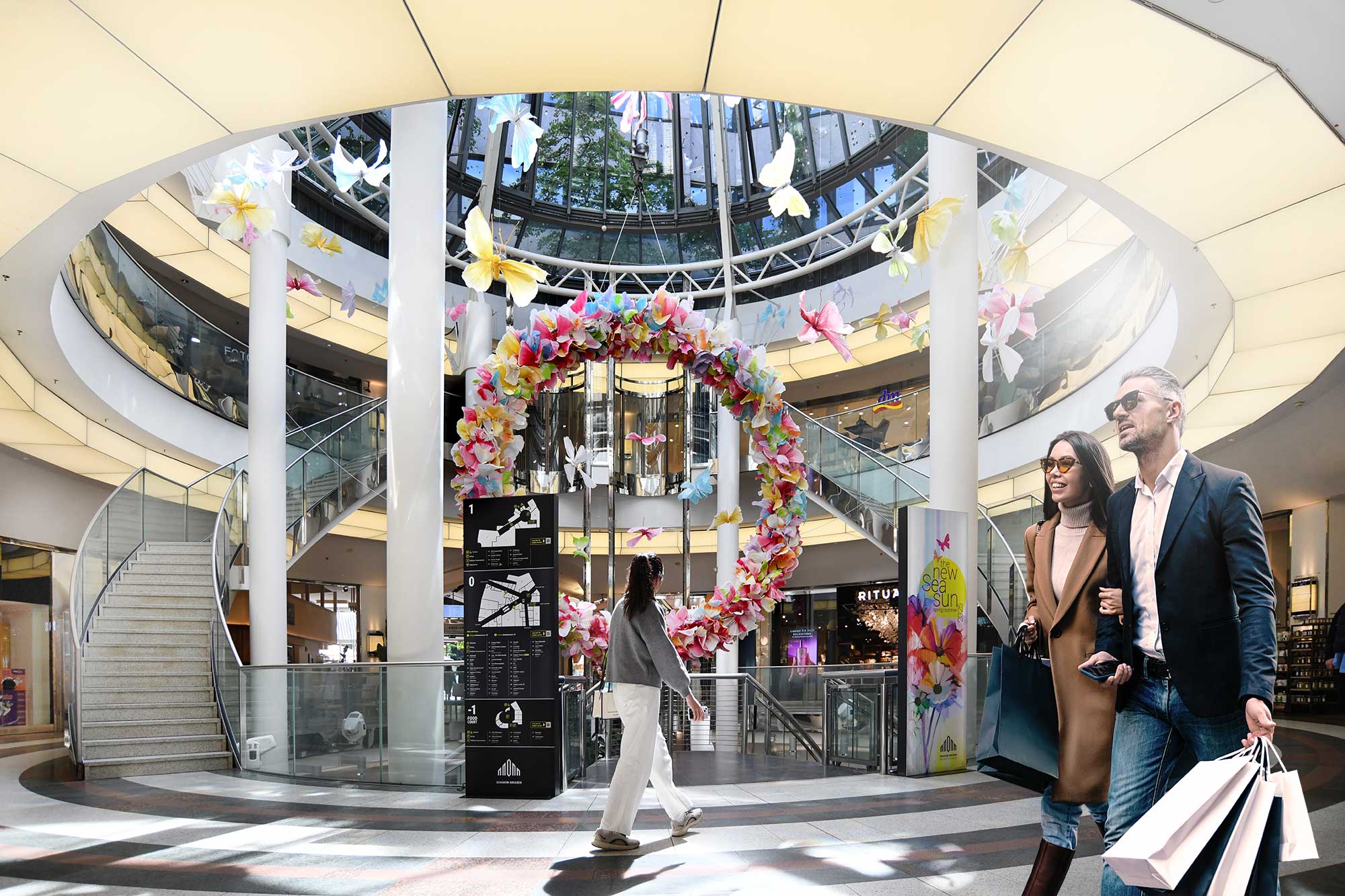 In the vibrant atrium of a shopping center, shoppers can admire colorful flowers and butterflies while exploring the space.