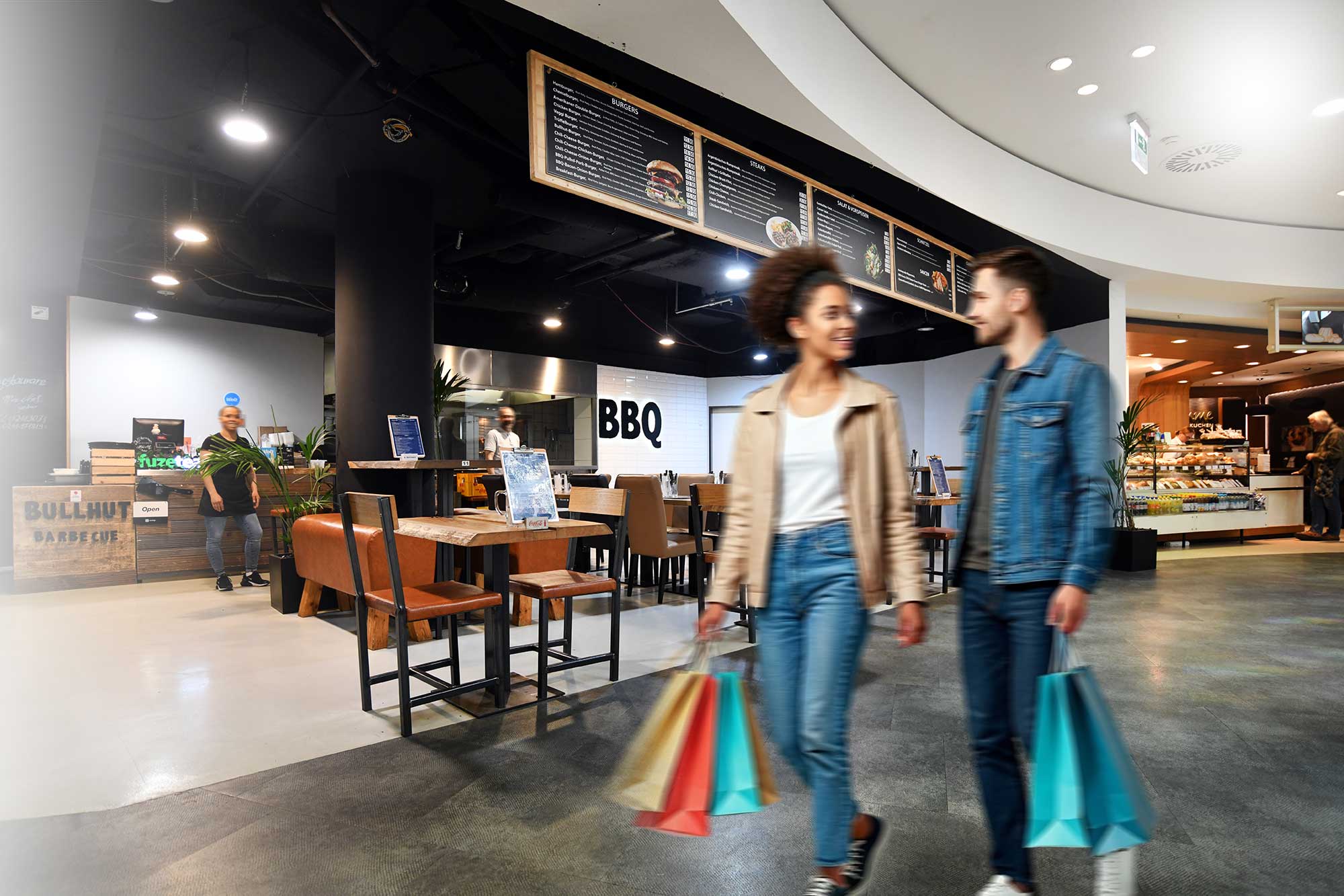 A lively food court scene with a grill stand, wooden tables, hanging menus, and shoppers carrying colorful bags.