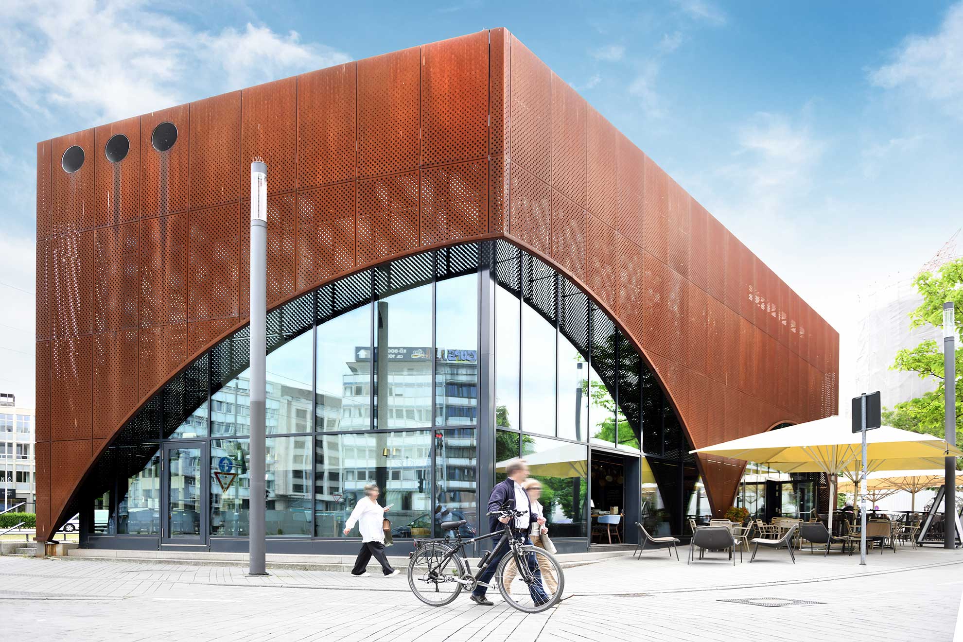 Modern architecture with a rust-colored facade featuring large glass windows and a bicycle parked in front, nestled in an urban environment.