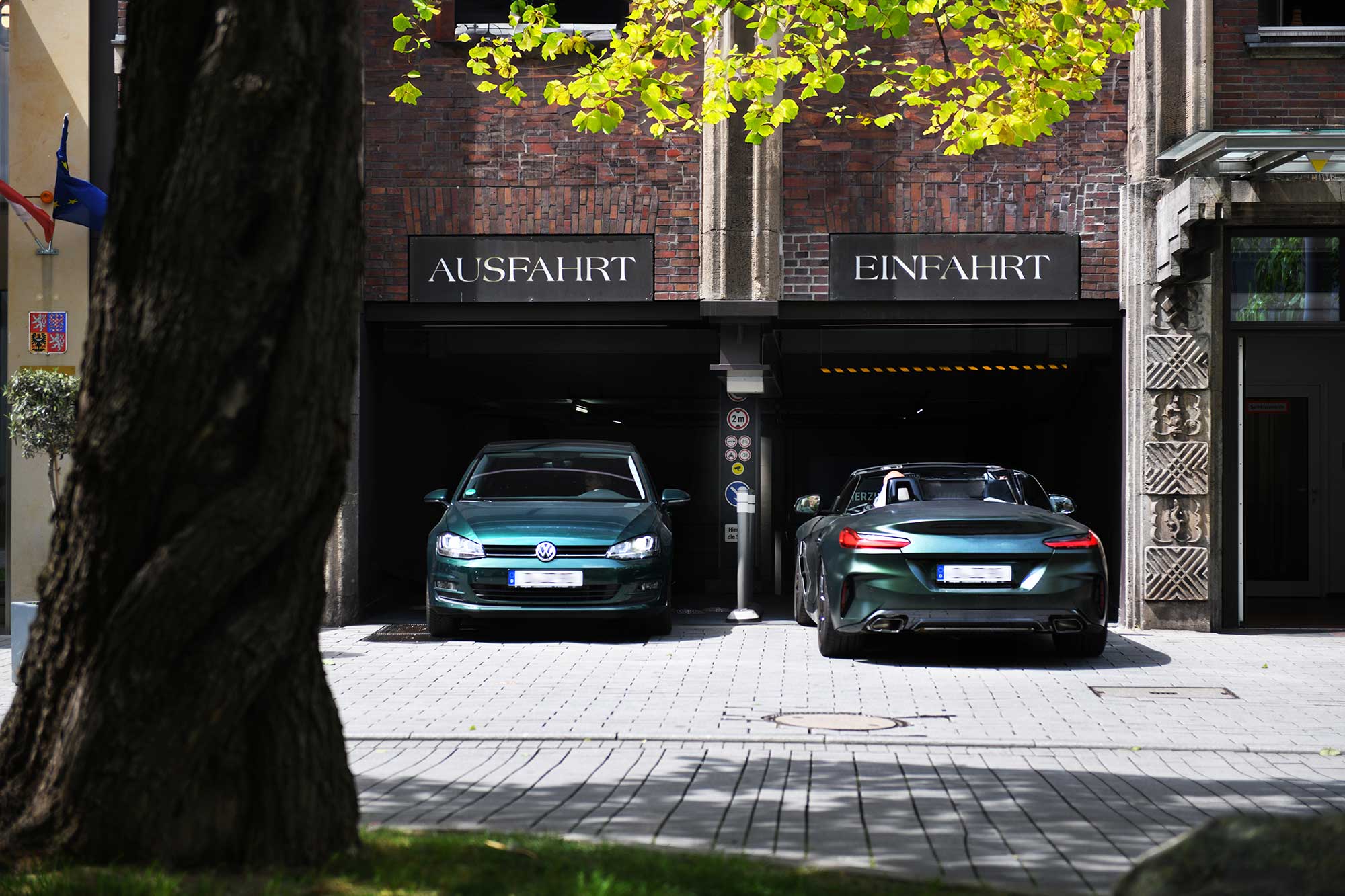 Two green cars, a Volkswagen and a sports car, parked in front of garage entrances marked “AUSFAHRT” and “EINFAHRT.”