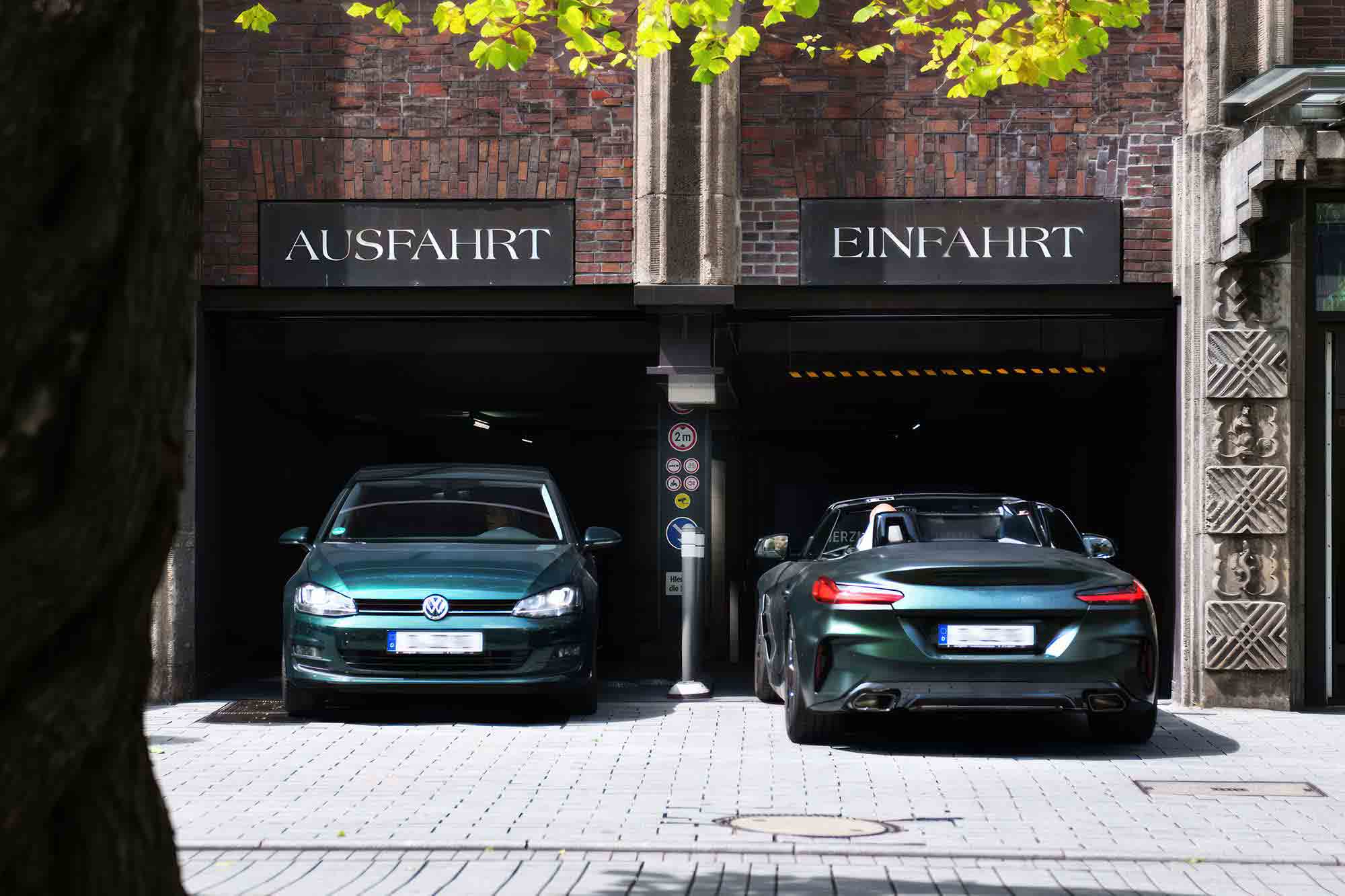 Two green cars, a Volkswagen and a sports car, parked in front of garage entrances labeled “AUSFAHRT” and “EINFAHRT.”
