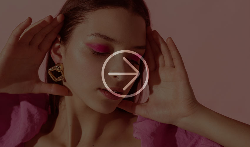 Young woman with pink eyeshadow and gold earrings against a pink background.