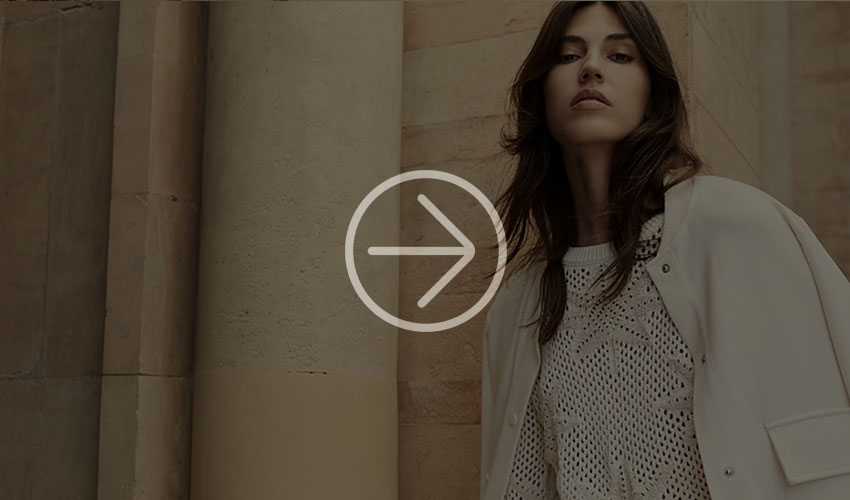 Woman in white clothing stands before stone columns, presenting a knitted sweater in a softly lit setting.