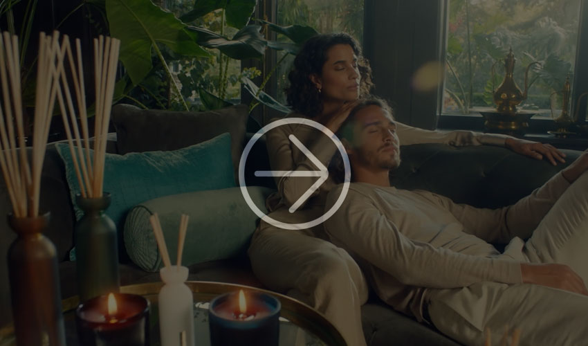 Woman sits on a sofa, placing her hand on a man's head, surrounded by scented candles, reed diffusers, and plants.