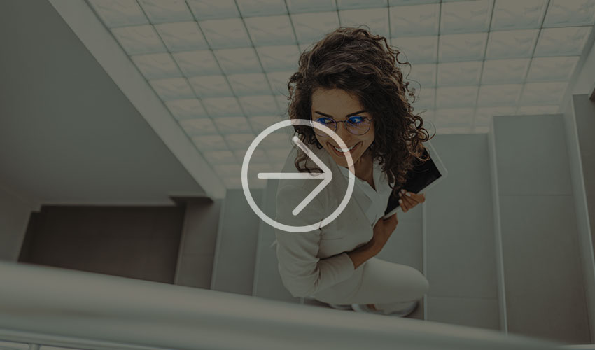 Woman in a white outfit climbs a modern staircase with a device in her hand, while natural light falls through a glass ceiling above.