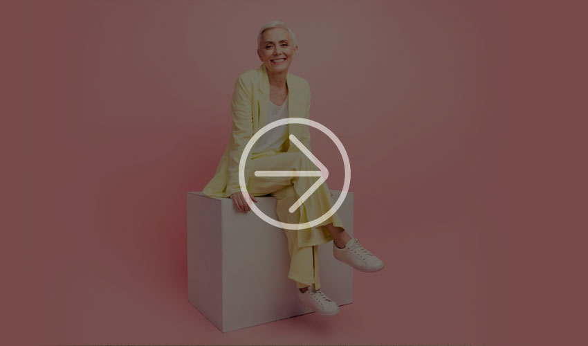 Person in a pastel yellow suit poses on a white cube against a soft pink background, wearing white sneakers.