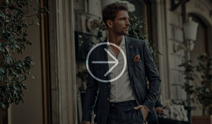 Man in a dark blue checked suit and white shirt stands confidently outside, surrounded by greenery and architecture.