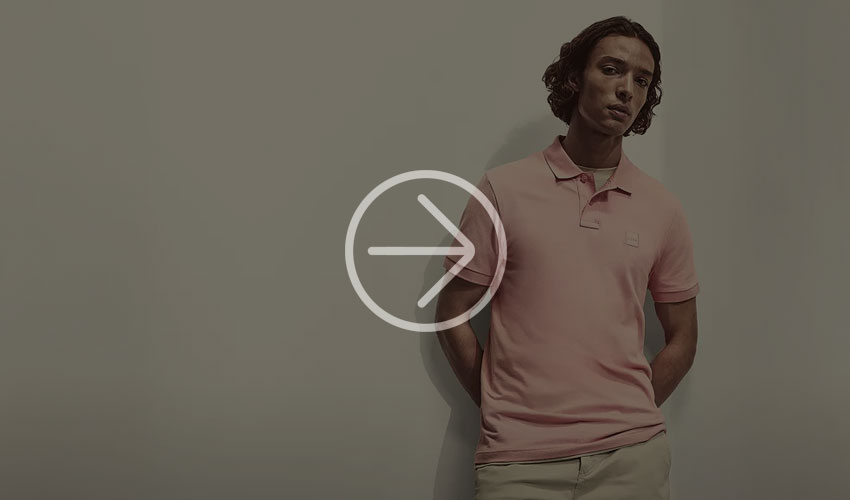 Man with longer hair stands in front of a wall wearing a pink T-shirt.