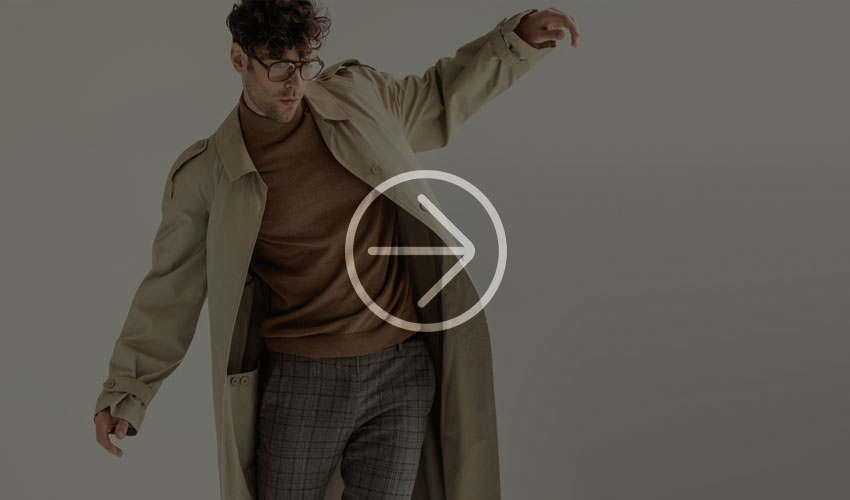 Man wearing a beige trench coat over a brown sweater and plaid trousers.