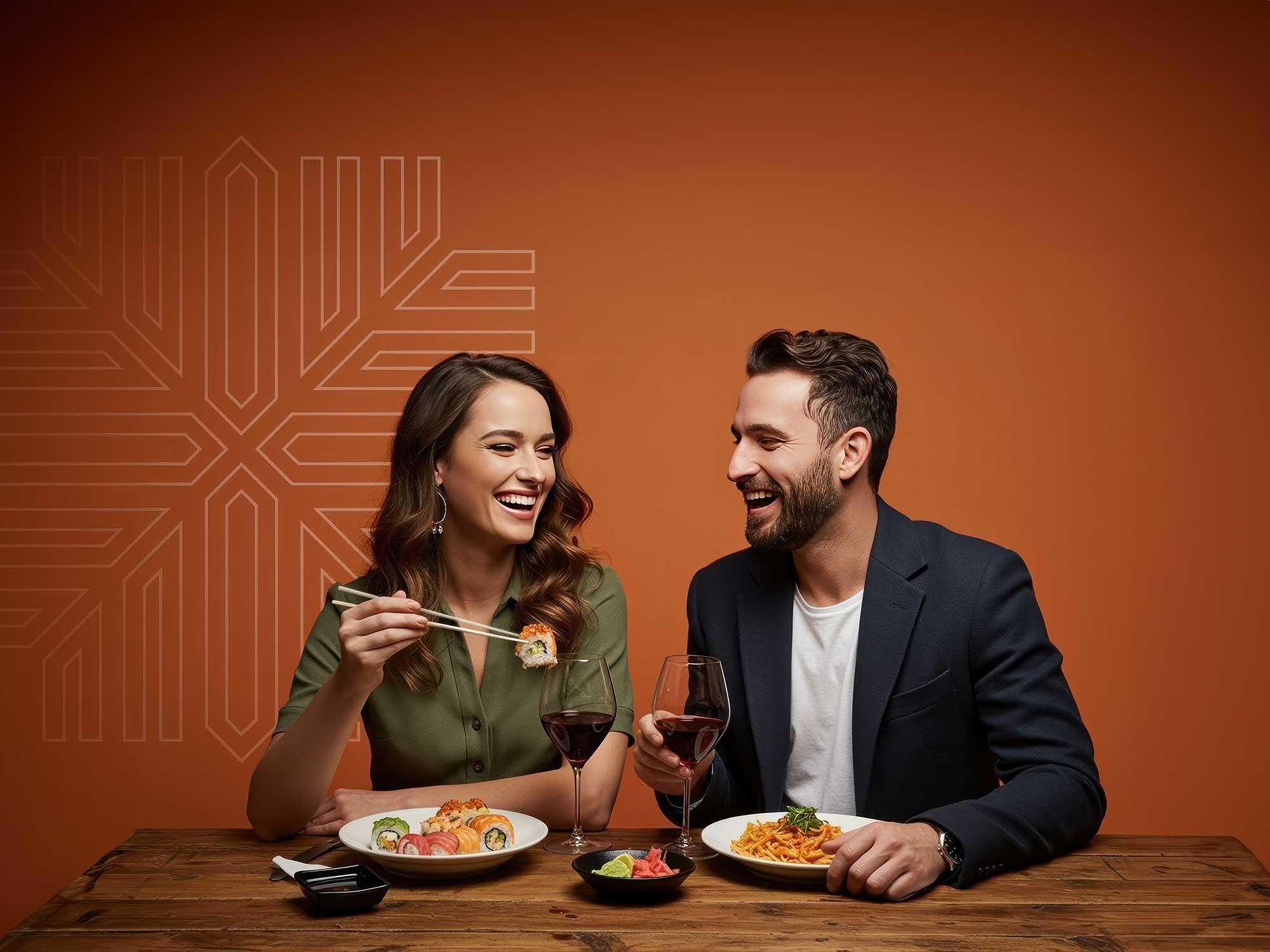 A couple enjoys a candlelit dinner with sushi, pasta, and red wine against a warm, orange background.