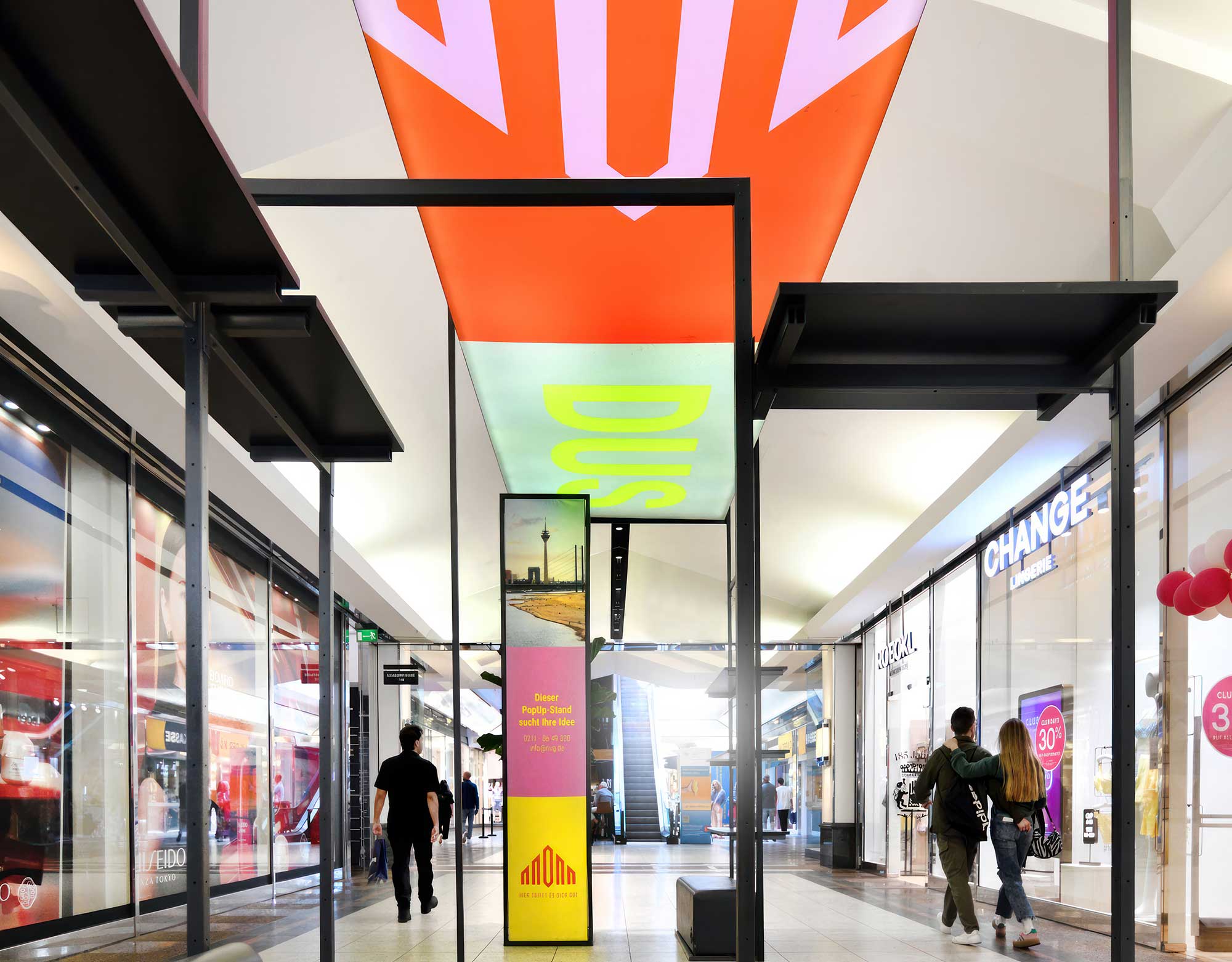 Ein belebter Korridor eines Einkaufszentrums mit bunten Schildern, vorbeigehenden Käufern und Schaufenstern auf beiden Seiten. Helle Lichter erhellen den Raum.