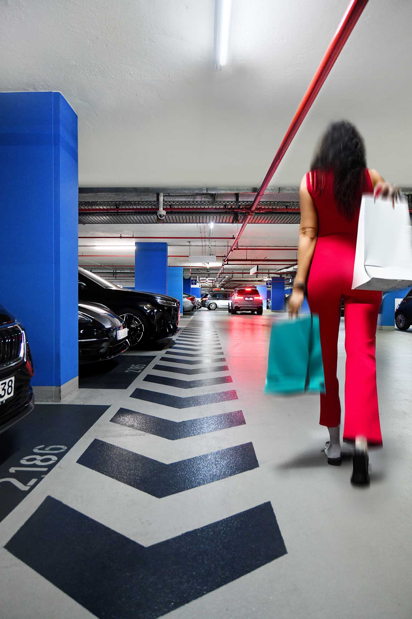 Eine Frau in einem roten Outfit geht mit blaugrünen und weißen Einkaufstüten durch ein hell erleuchtetes Parkhaus.