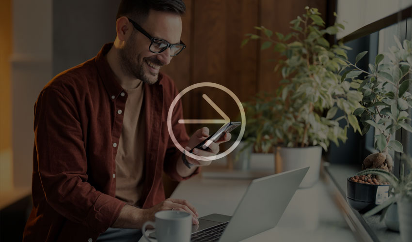 Lächelnder Mann mit Brille arbeitet am Laptop, schaut auf sein Smartphone, mit Kaffeetasse und Pflanzen am Fenster.