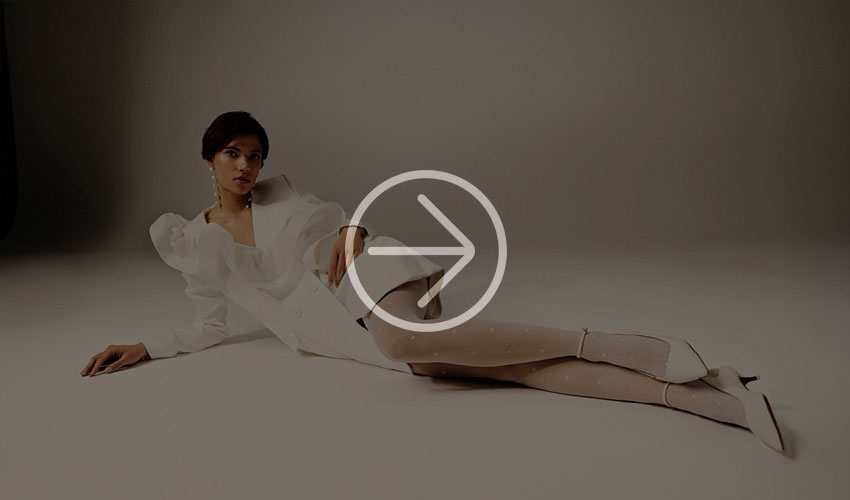 Model in weißem Outfit mit gepunkteten Strumpfhosen und weißen Schuhen, anmutig vor sanftem Hintergrund.       