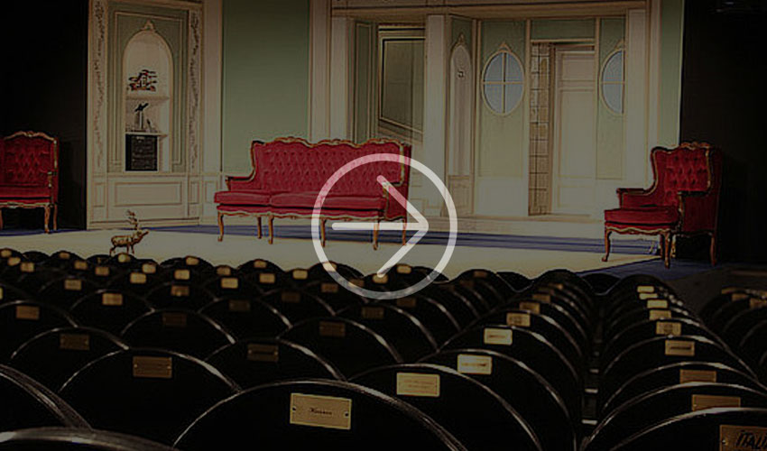 Theaterbühne mit roten Samtstühlen vor dekorativem Hintergrund und schwarzen Sitzreihen.