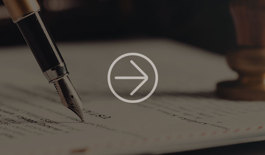 Close-up of a fountain pen hovering over a document. A blurred wooden stamp can be seen in the background, suggesting a formal handwriting or signature.