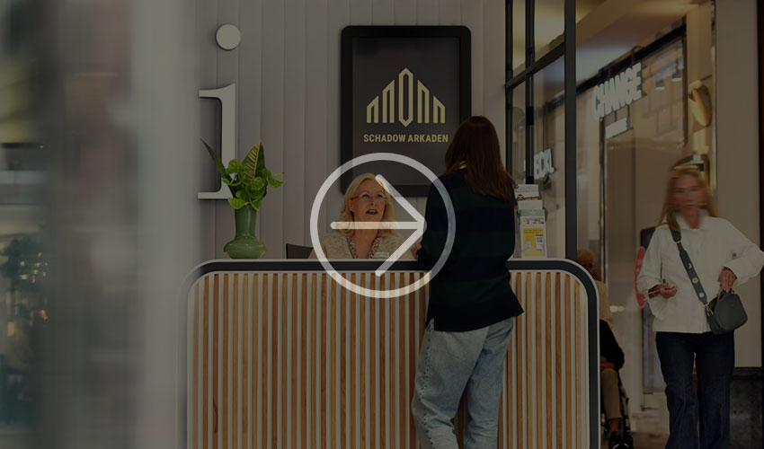 A wooden reception counter in a shopping center with a plant and an information sign. Shoppers can be seen in the background.