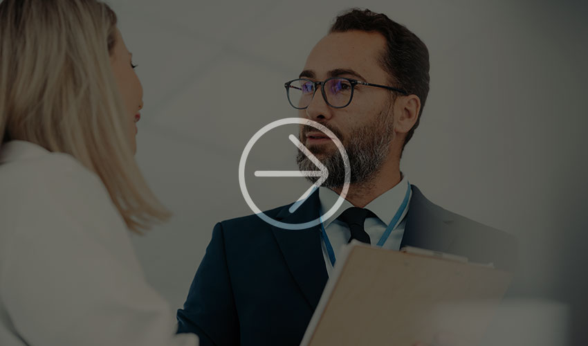 A professional woman in a white blazer talks to a man in a suit in a bright office, who is holding a clipboard in his hand.